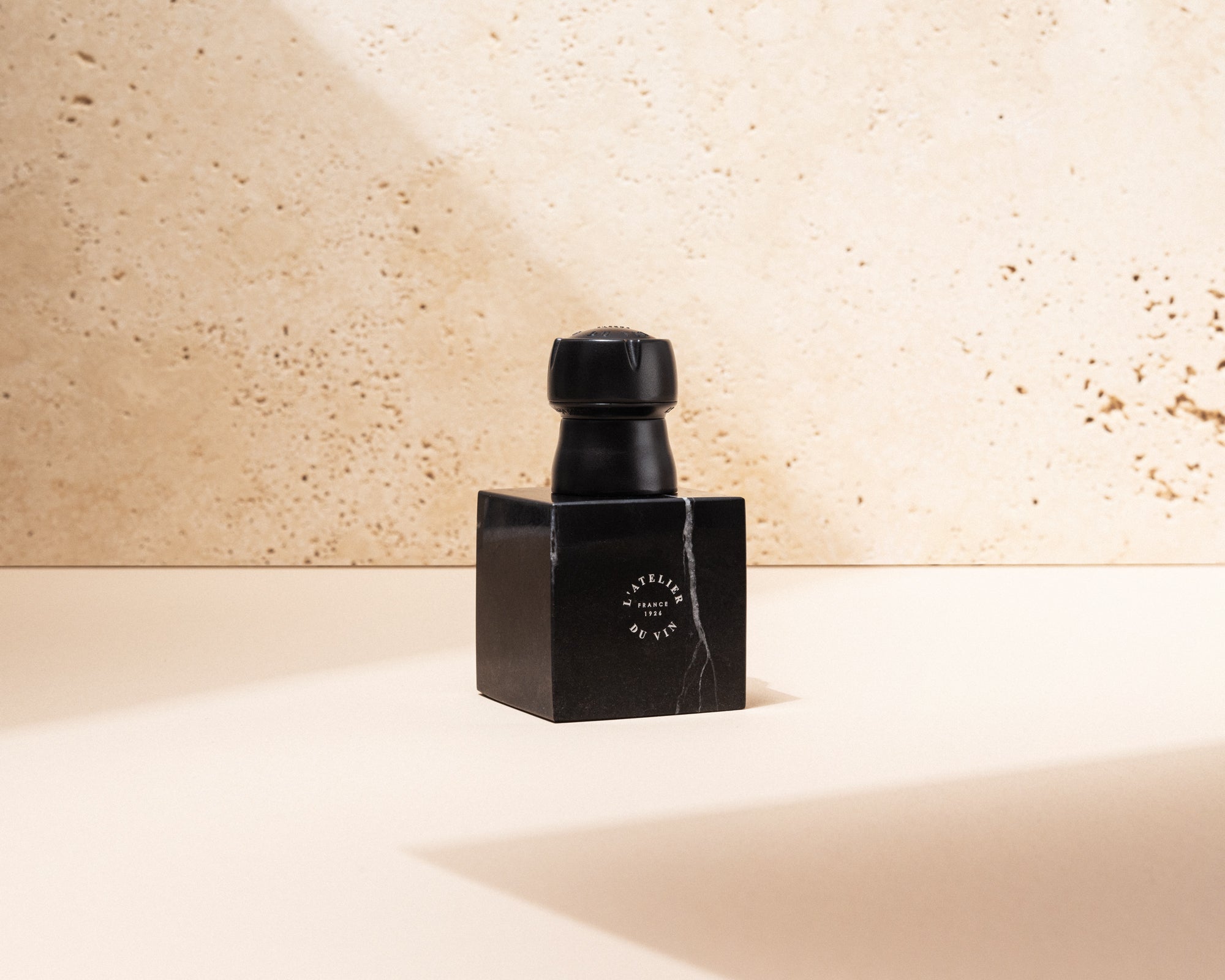 L'Atelier du Vin Bubble Cork Black sobre cubo de mármol en superficie beige con sombra diagonal