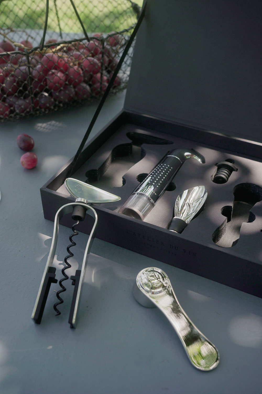 Set L'Atelier du Vin Oeno Collection 2 con accesorios sobre mesa oscura junto a racimo de uvas