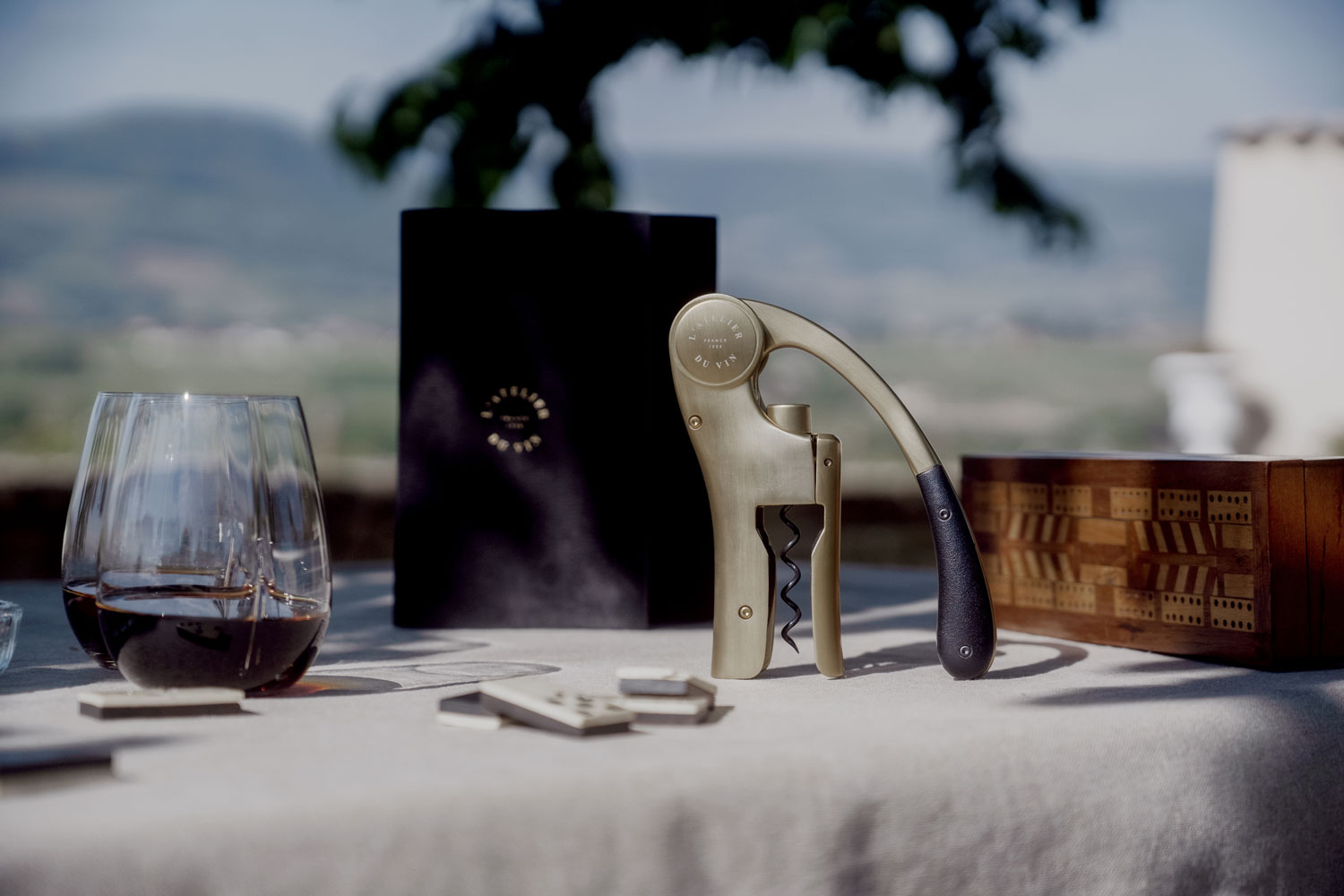 Sacacorchos Oeno Motion Gold Nomad L'Atelier du Vin en mesa exterior con copa de vino tinto y estuche