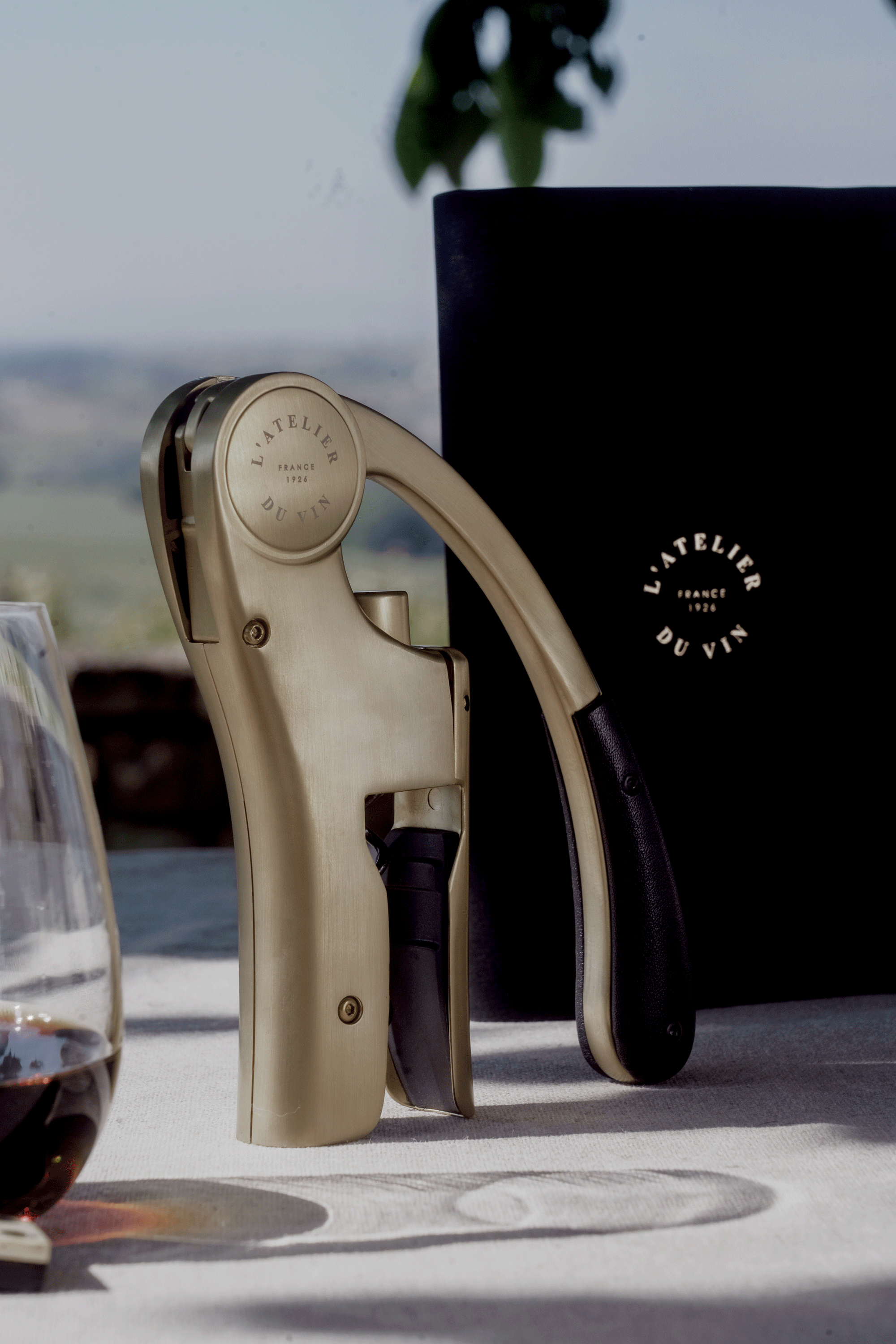 Sacacorchos Oeno Motion Gold Nomad L'Atelier du Vin en exterior con copa de vino y estuche negro al fondo