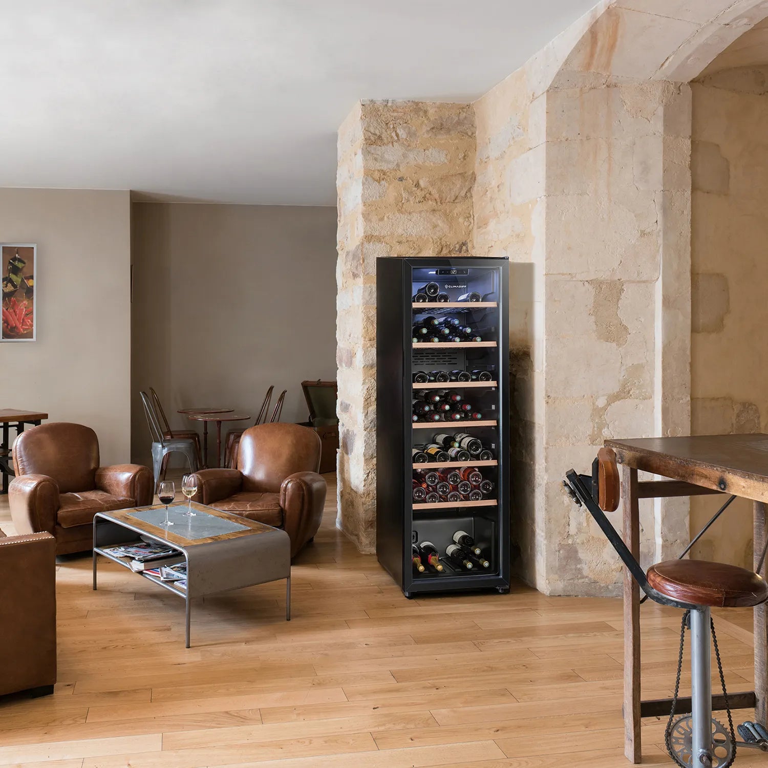 Vinoteca Climadiff CPW147 colocada contra una pared de piedra en una sala de estar elegante, decorada con sillones de cuero marrón, una mesa baja y suelos de madera clara.
