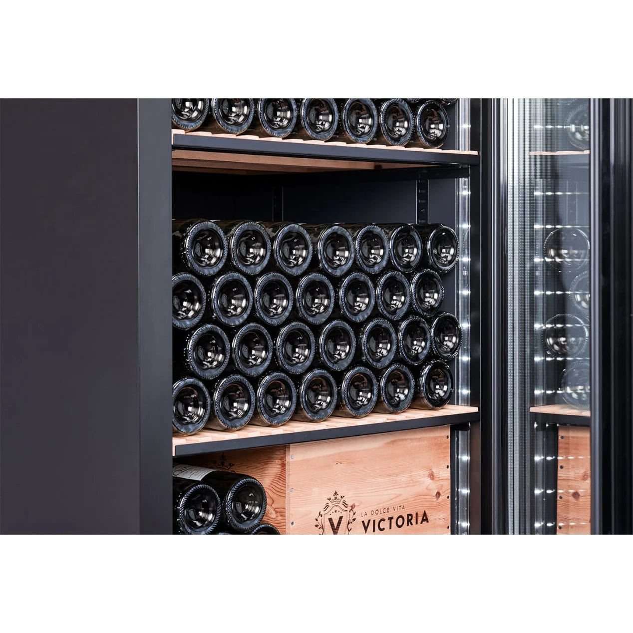 Vista lateral cercana del interior de la vinoteca mQuvée VELVET 220 Premium Package, mostrando varias botellas de vino apiladas horizontalmente en estantes de madera, con una caja de madera con el texto “VICTORIA” en la parte inferior y luces LED verticales encendidas en el marco.