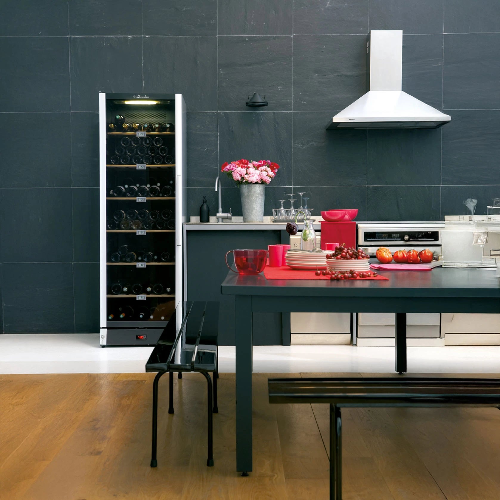 Vinoteca La Sommelière VIP185 colocada en una cocina moderna con paredes de pizarra, al lado de una encimera con fregadero, flores y utensilios de cocina, junto a una mesa con platos, copas y frutas.