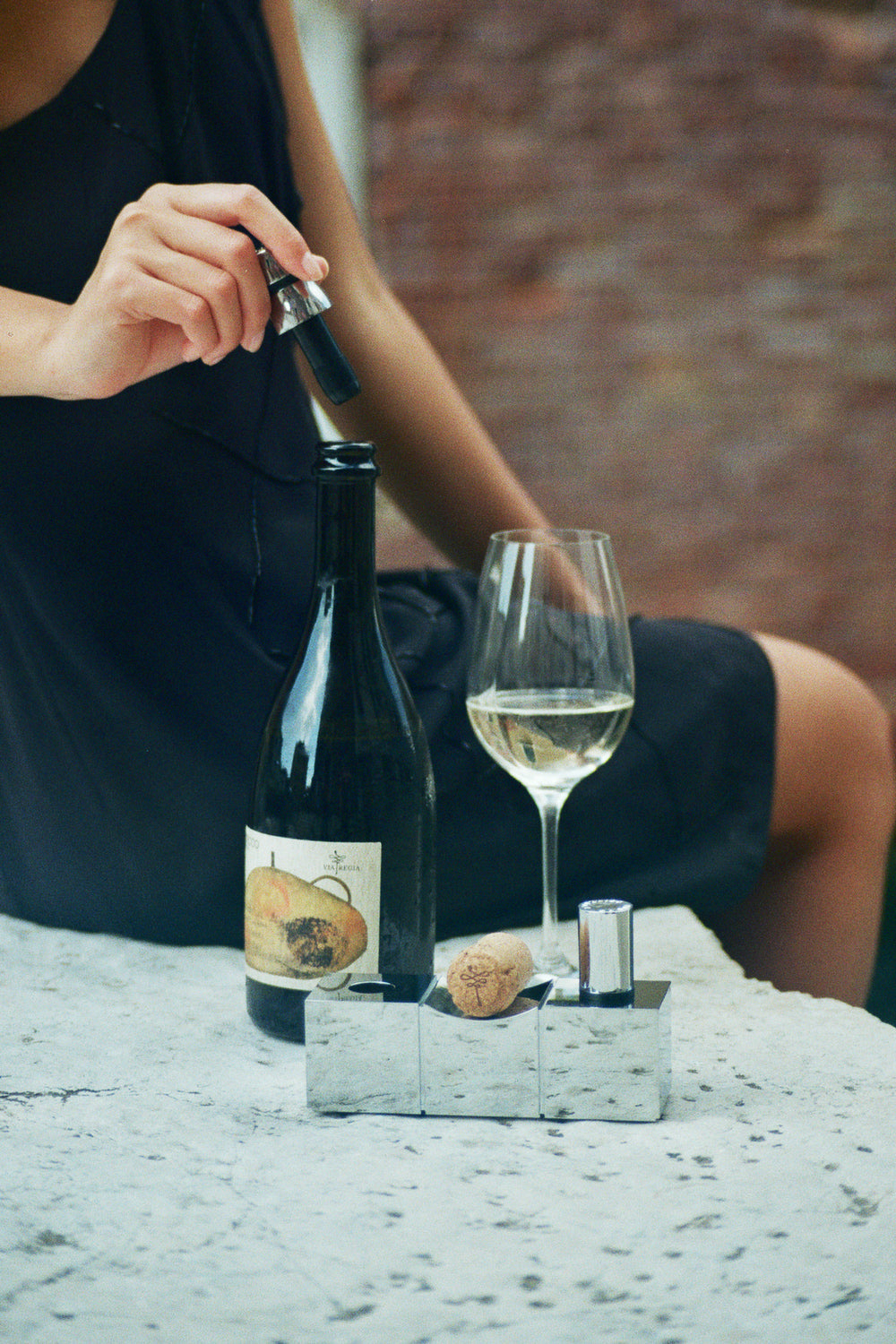 Mujer usando tapón de vino L'Atelier du Vin Bouchons Millésimes sobre mesa de mármol con copa de vino blanco