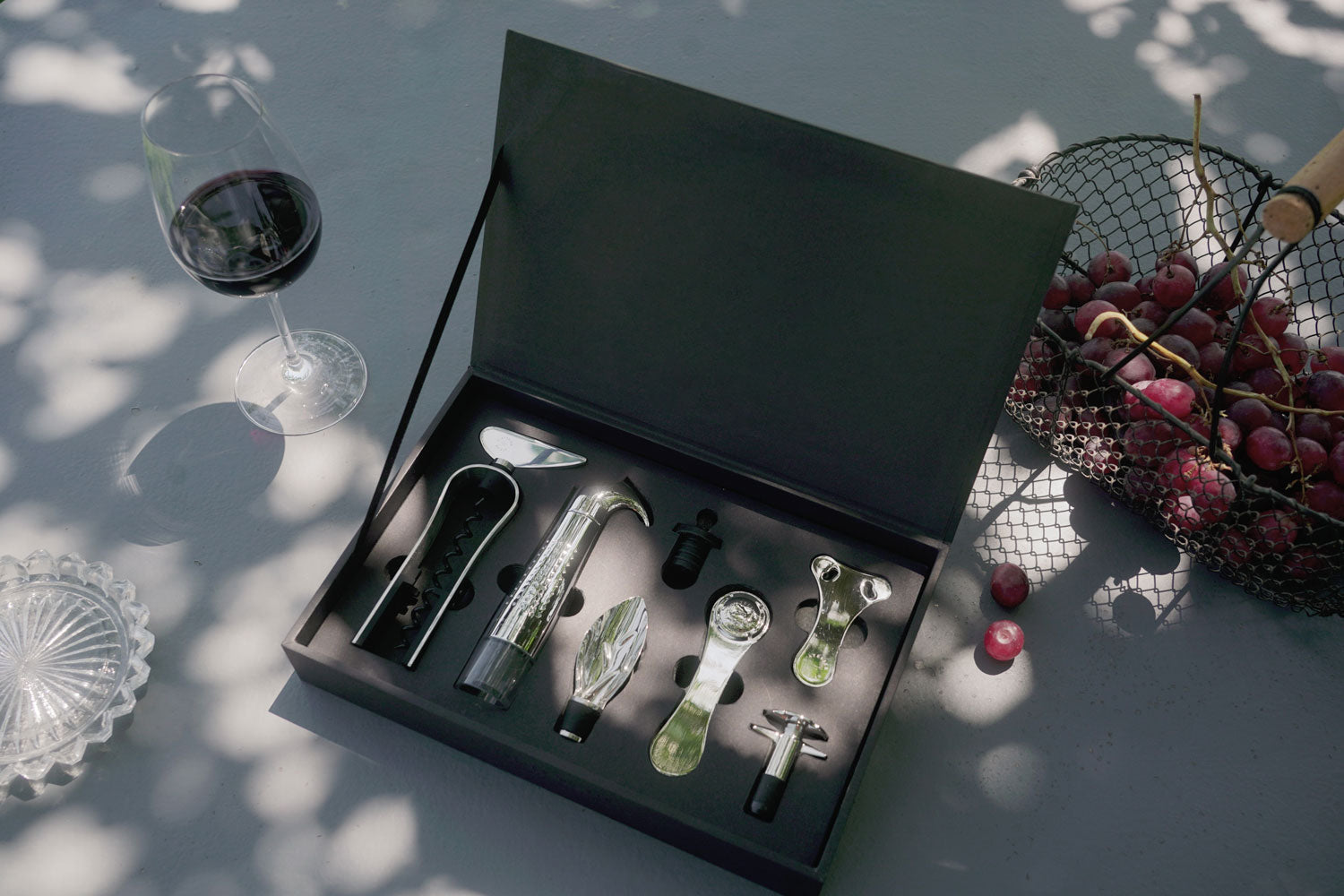 Set L'Atelier du Vin Oeno Collection 2 en terraza soleada con copa de vino tinto y cesta de uvas