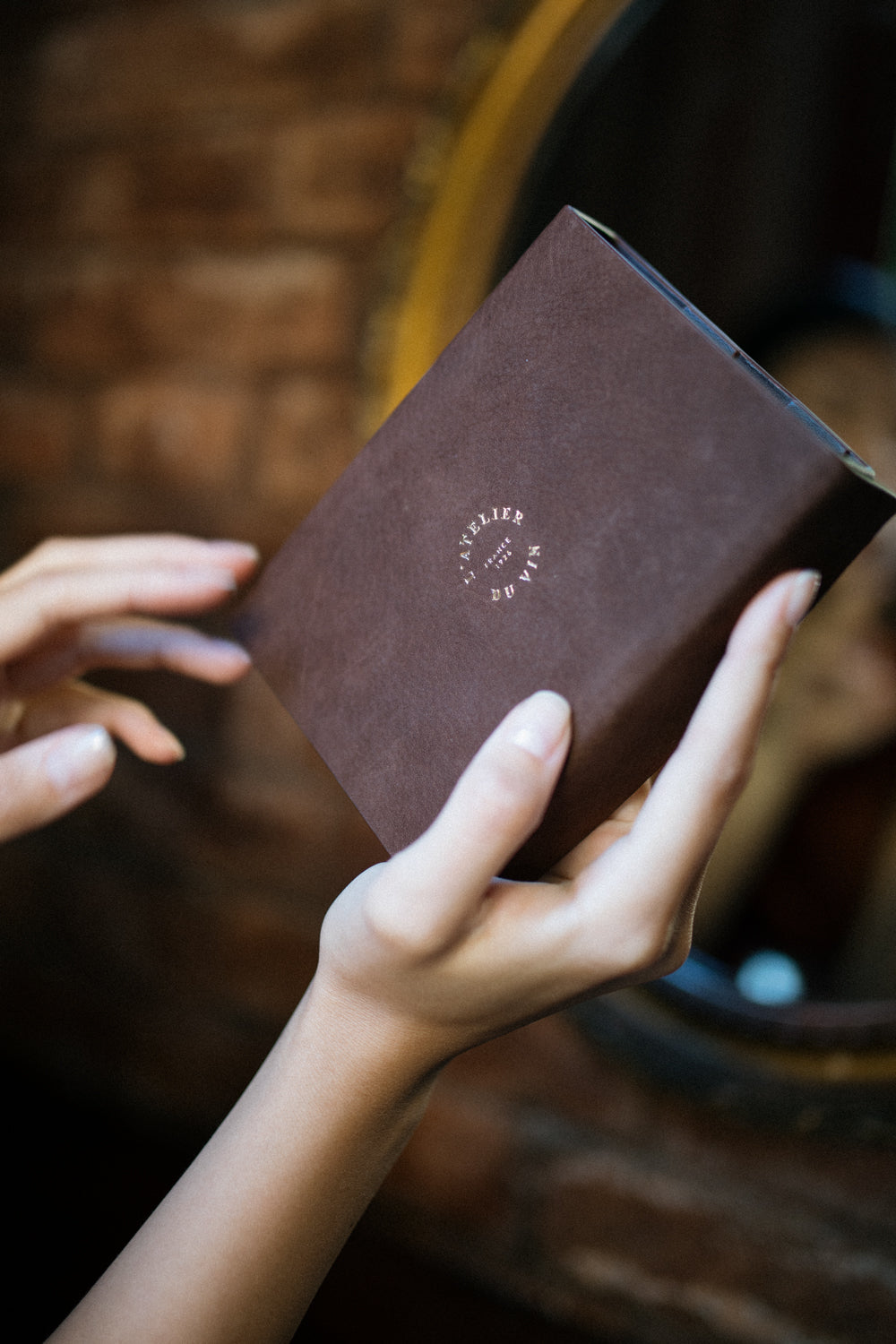 Manos sosteniendo el estuche de cuero marrón del set Les Bijoux de L'Atelier du Vin en ambiente cálido