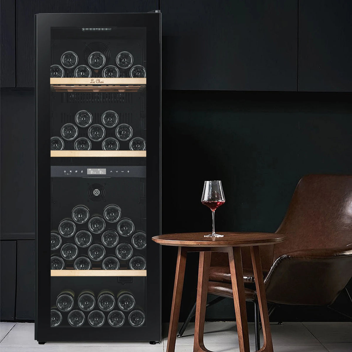 Vinoteca Le Chai LCT2FS100 de 100 botellas, en un salón junto a una mesa con una copa de vino tinto, con los estantes de madera sin frontal.