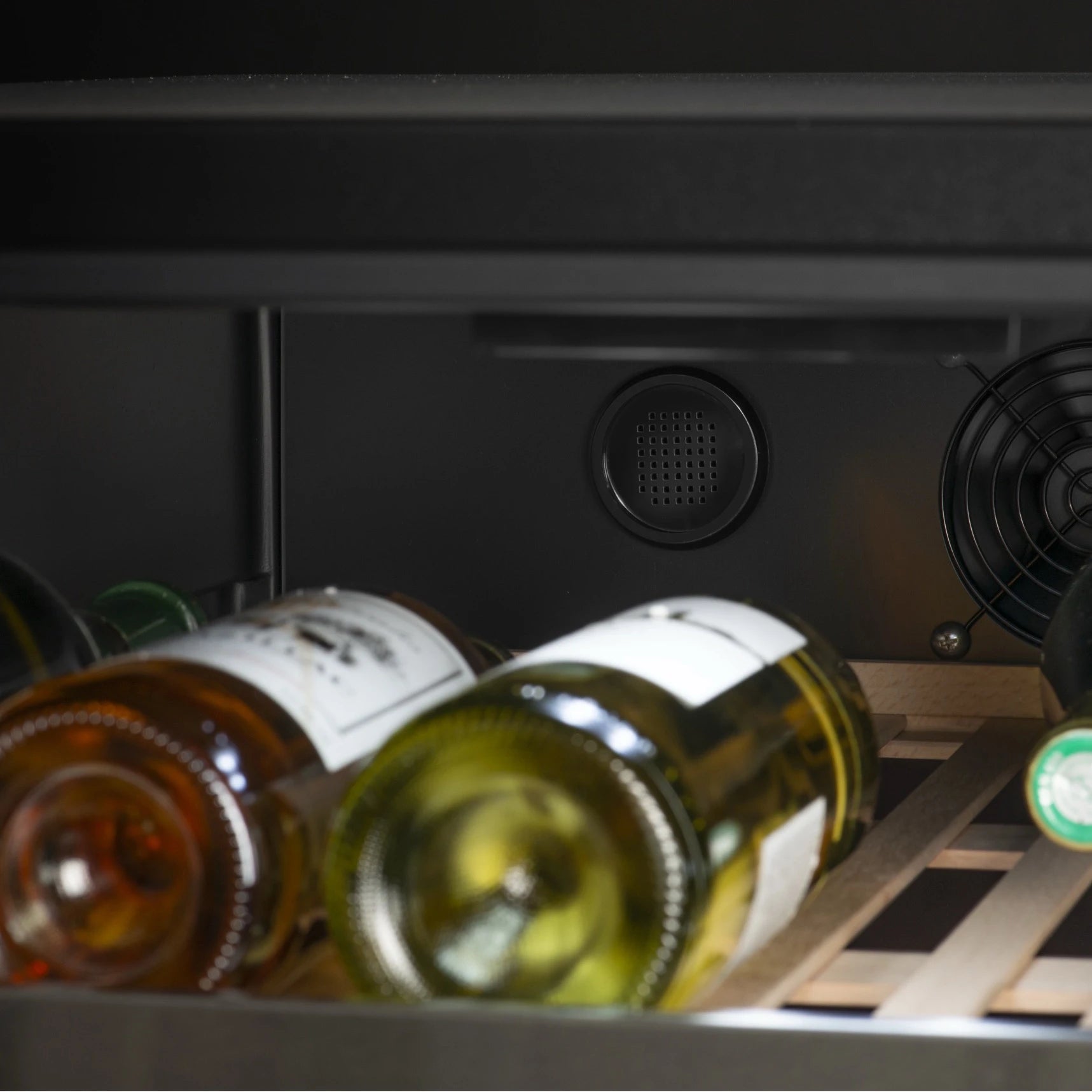 Primer plano del interior de la vinoteca La Sommelière MT150DZ, enfocando botellas de vino blanco colocadas sobre un estante de madera y el sistema de ventilación en el fondo, que incluye una rejilla circular y un ventilador negro lateral.