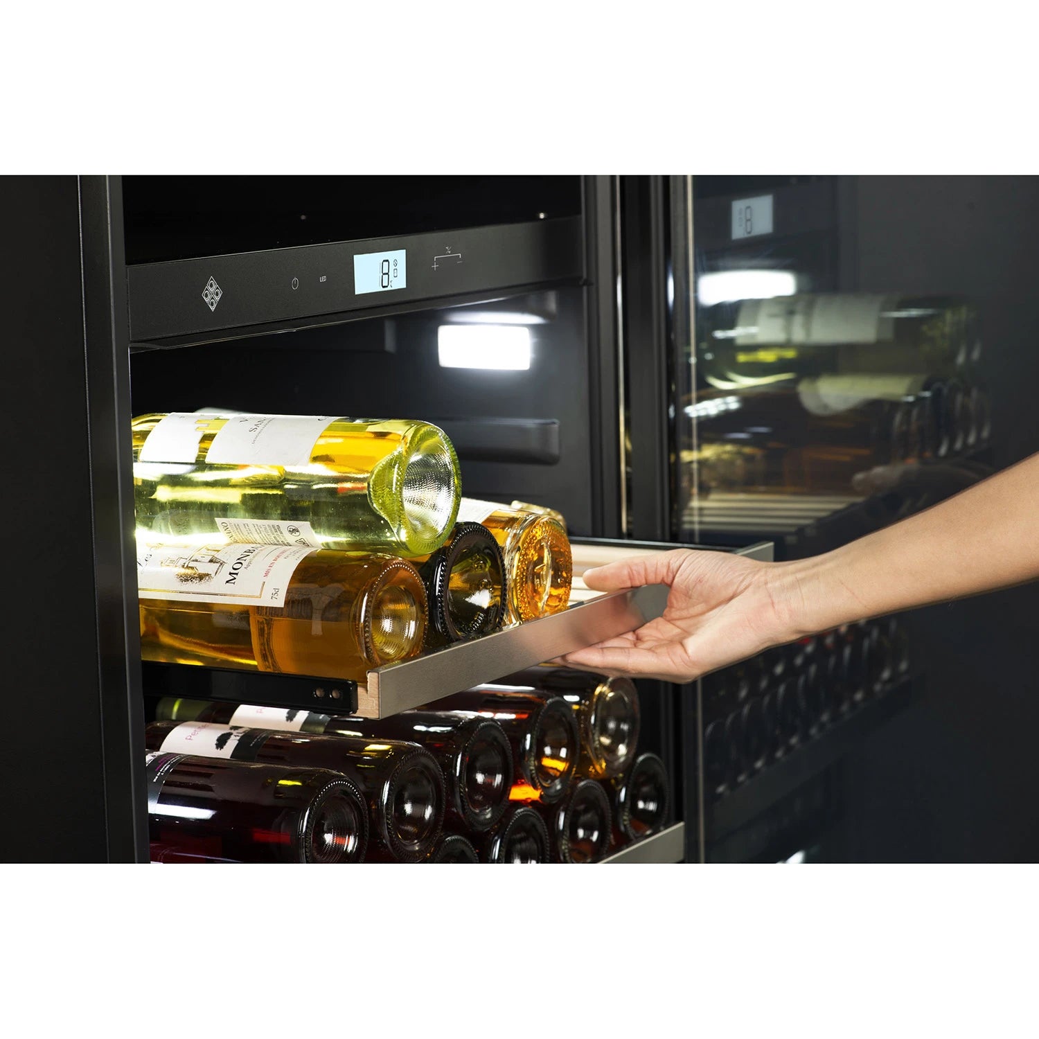 Mano abriendo un estante extraíble de la vinoteca La Sommelière MT150DZ, mostrando botellas de vino blanco almacenadas horizontalmente. En el fondo se observa una pantalla digital con temperatura de 8 °C y luz LED interior iluminando el compartimento.