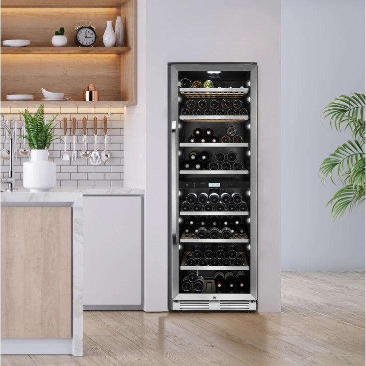 Vinoteca empotrada La Sommelière MT150DZ con capacidad para 152 botellas, instalada en una cocina moderna con acabados en madera clara y encimeras blancas. El electrodoméstico tiene puerta de vidrio, estantes de metal y doble zona de temperatura visible en el panel digital. Clasificación energética G, consumo de 181 kWh al año y nivel de ruido de 39 dB.