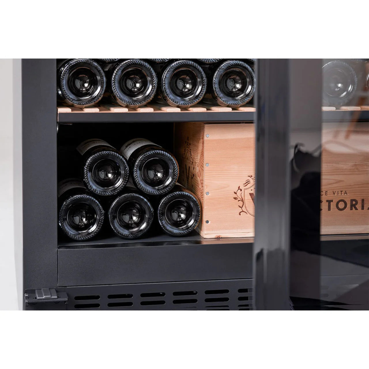 Detalle de la parte inferior de la vinoteca mQuvée, con botellas de vino apiladas horizontalmente junto a una caja de madera decorativa con la inscripción “Victoria”.