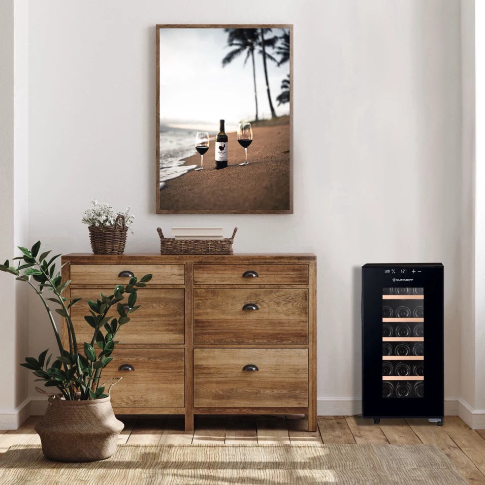 Vinoteca Climadiff CUVEE18 colocada junto a un aparador de madera en una sala con decoración natural, acompañada de plantas y un cuadro de playa en la pared.