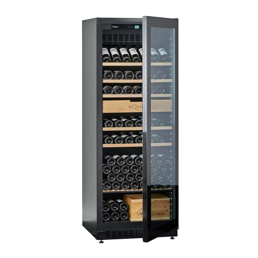 Vista en ángulo de la vinoteca mQuvée VELVET 220 Premium Package con la puerta de vidrio parcialmente abierta, mostrando varias botellas de vino almacenadas en estantes de madera y dos cajas de madera con logotipos visibles.