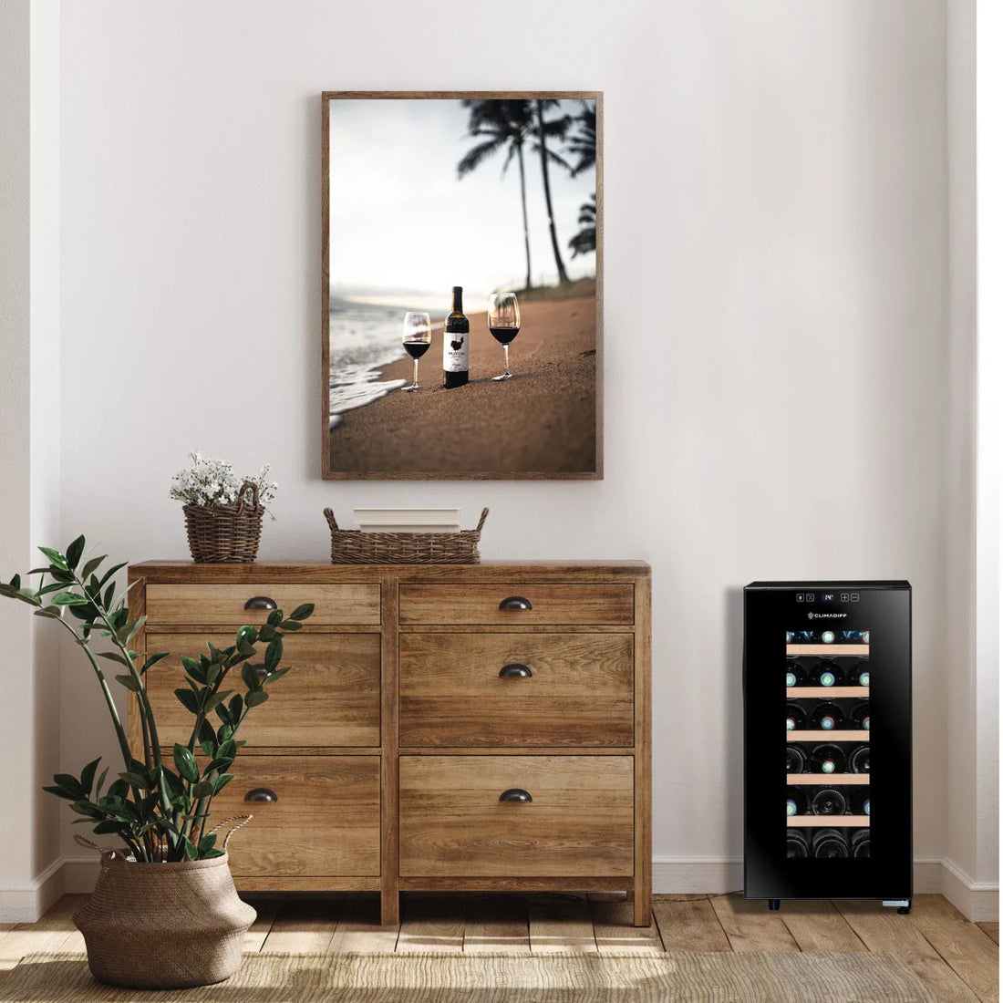 Vinoteca pequeña de diseño moderno junto a una cómoda de madera clara. Decoración incluye planta en maceta y cuadro con vino en la playa.
